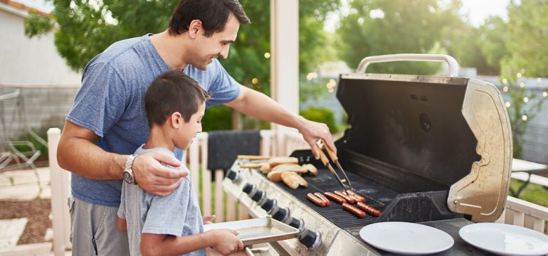A father teaching his son grilling techniques on a backyard gas grill.