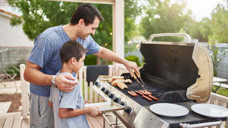 A father teaching his son grilling techniques on a backyard gas grill.