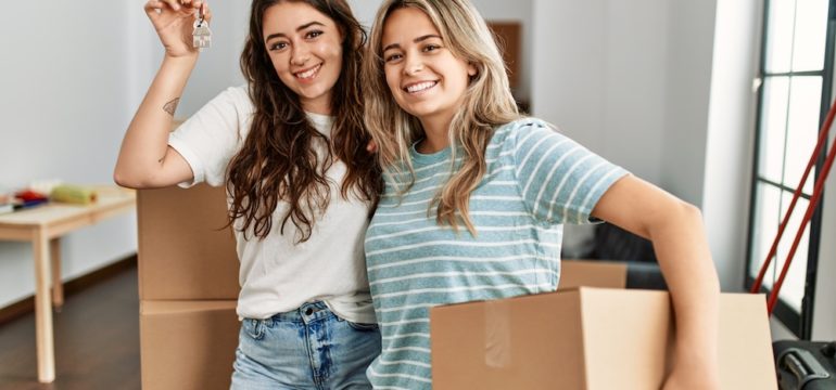 Two smiling ladies holding a cardboard box and key to show buy a house with a friend.