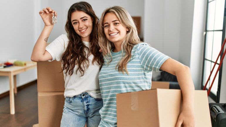 Two smiling ladies holding a cardboard box and key to show buy a house with a friend.