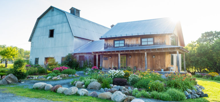 Photo of a unique home that is an old farmhouse with a barn attached and beautiful flower gardens in the morning sun.