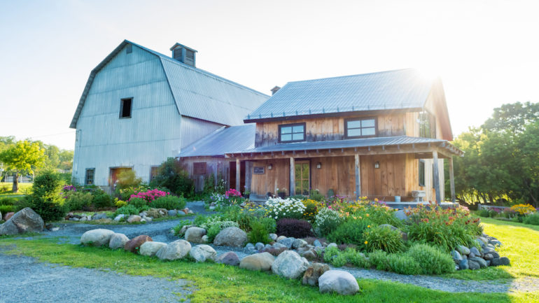 Photo of a unique home that is an old farmhouse with a barn attached and beautiful flower gardens in the morning sun.