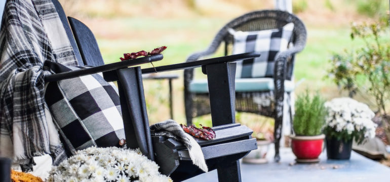 Adirondack rocking chair with traditional style buffalo check blanket and pillows on a front porch decorated for autumn with heirloom gourds and white and orange mums.