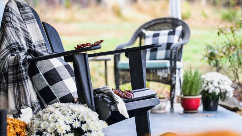 Adirondack rocking chair with traditional style buffalo check blanket and pillows on a front porch decorated for autumn with heirloom gourds and white and orange mums.