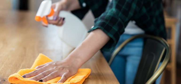 Close-up view of a woman wiping dust using spray and an orange microfiber cleaning cloth on the dirty table.