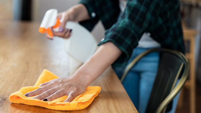 Close-up view of a woman wiping dust using spray and an orange microfiber cleaning cloth on the dirty table.