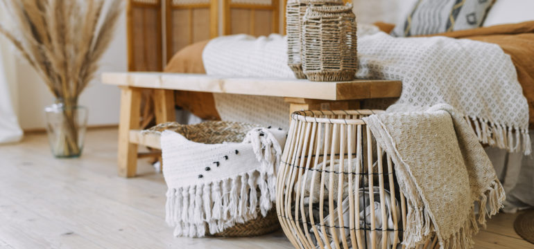 Post-pandemic interior design trends are shown in Boho design. Comfortable bedroom with textile sheet on the bed, wooden bench seat, bamboo dressing screen, dry plants in a vase, and wicker basket.