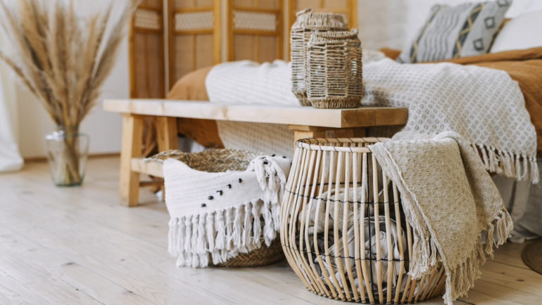 Post-pandemic interior design trends are shown in Boho design. Comfortable bedroom with textile sheet on the bed, wooden bench seat, bamboo dressing screen, dry plants in a vase, and wicker basket.