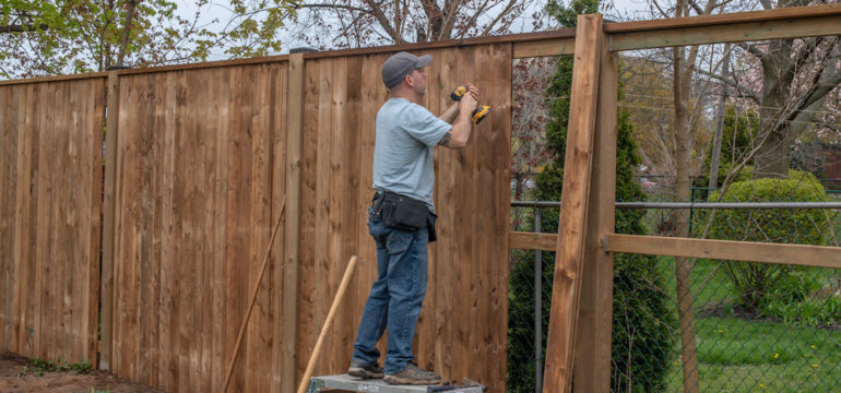 A white, middle-aged man builds a wooden fence in his backyard.