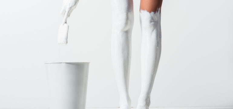 Cropped image of woman with legs painted with white paint holding brush above bucket on the white floor.
