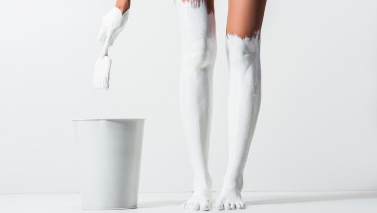 Cropped image of woman with legs painted with white paint holding brush above bucket on the white floor.