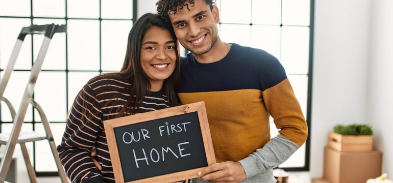 Young Latin couple smiling happily, holding "our first home" written on a blackboard in their new starter home.
