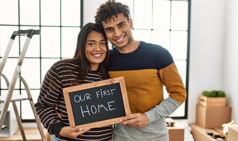 Young Latin couple smiling happily, holding "our first home" written on a blackboard in their new starter home.