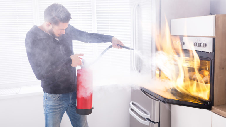 Man using red fire extinguishers to stop a fire in a home kitchen oven.