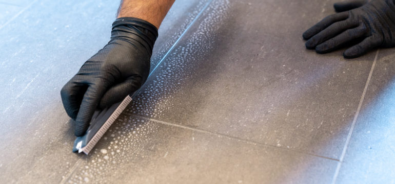 A close-up of a professional cleaner cleaning dirty tile grout with a brush blade and foamy soap on a gray bathroom floor.