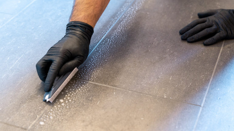 A close-up of a professional cleaner cleaning dirty tile grout with a brush blade and foamy soap on a gray bathroom floor.