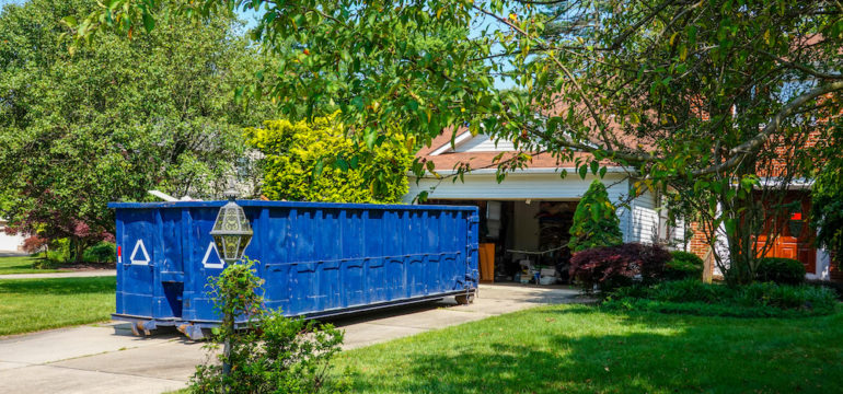 An empty blue dumpster in the driveway of a house with its garage door open in a residential community.