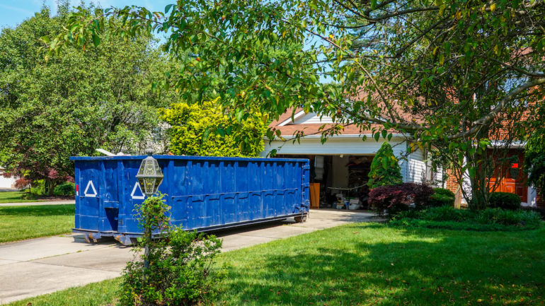 An empty blue dumpster in the driveway of a house with its garage door open in a residential community.