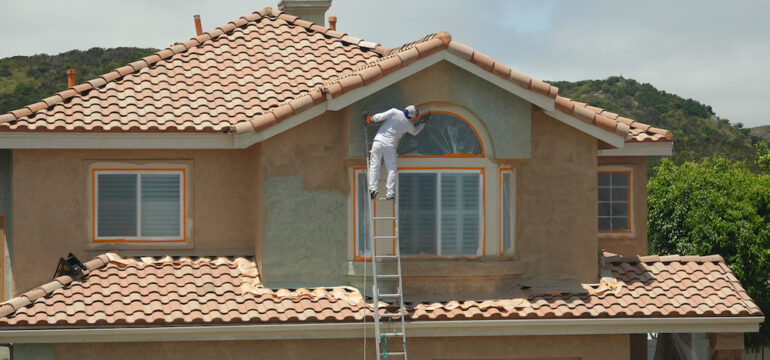 Worker leaning dangerously off the ladder to paint the stucco on a suburban home.
