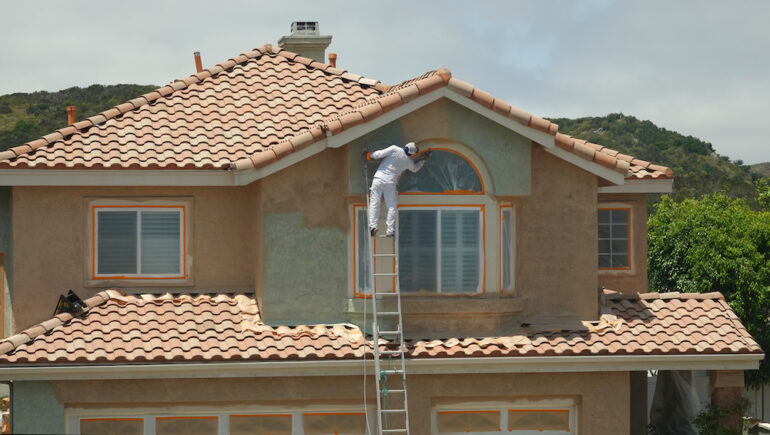 Worker leaning dangerously off the ladder to paint the stucco on a suburban home.