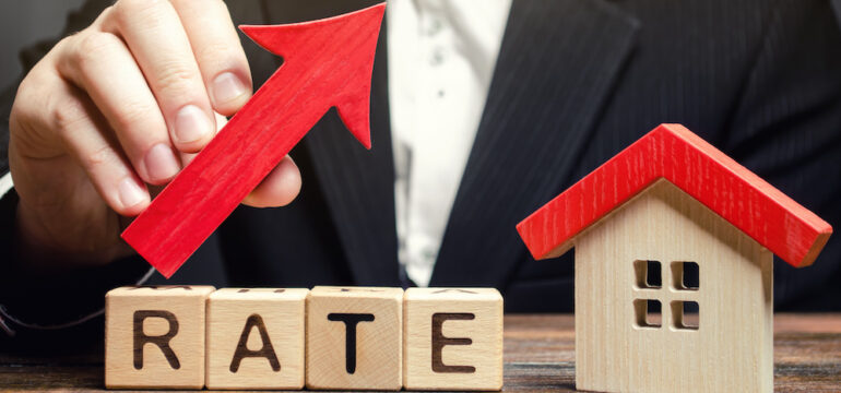 A man holds a red arrow up above the word Rate and a wooden house. The concept of raising interest rates calls for creative mortgages as a solution.