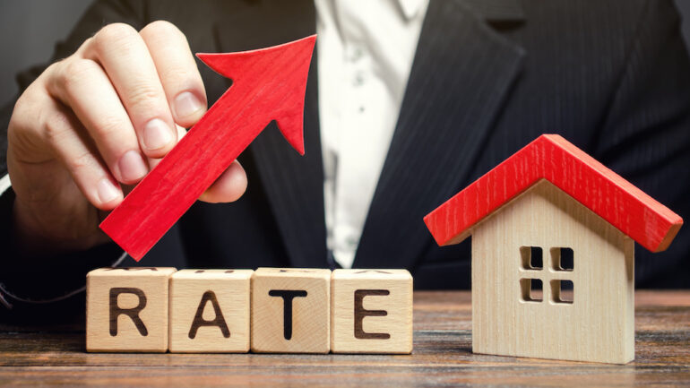 A man holds a red arrow up above the word Rate and a wooden house. The concept of raising interest rates calls for creative mortgages as a solution.