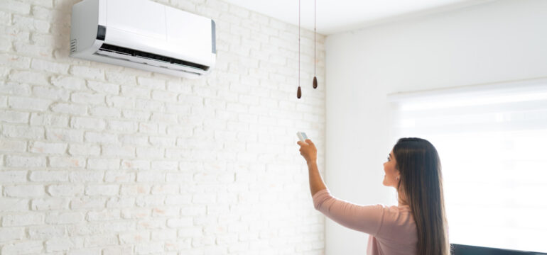 Young woman adjusting temperature of a mini-split air conditioner using remote control in a room at home.