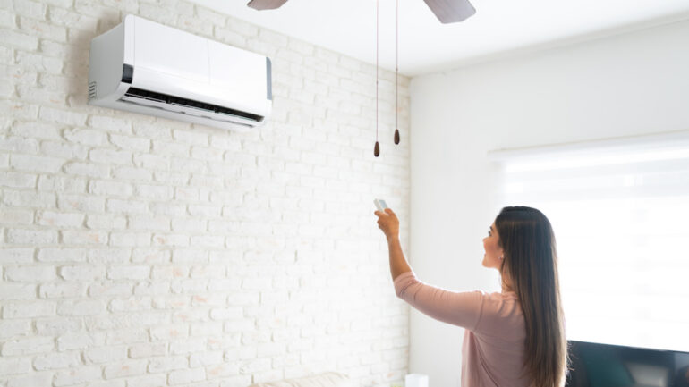 Young woman adjusting temperature of a mini-split air conditioner using remote control in a room at home.