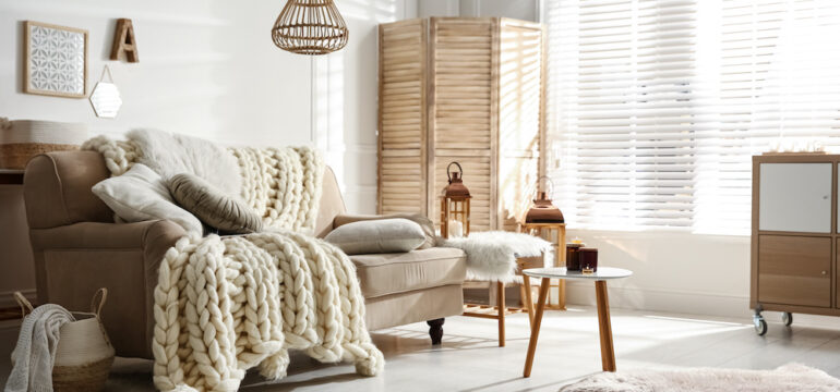 Cozy living room interior with beige sofa, knitted blankets, cushions, and off-white window blinds and shades.