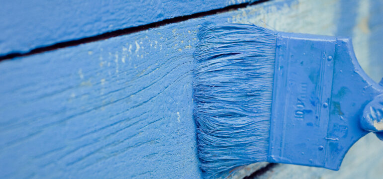 Blue ceramic house paint is applied with a paintbrush on an exterior shingle.
