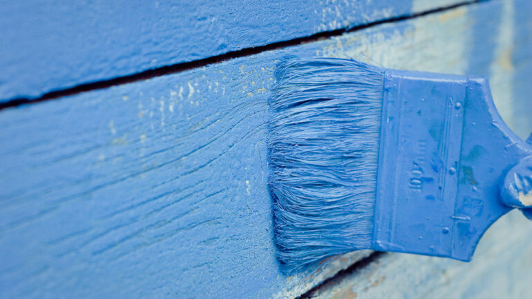 Blue ceramic house paint is applied with a paintbrush on an exterior shingle.