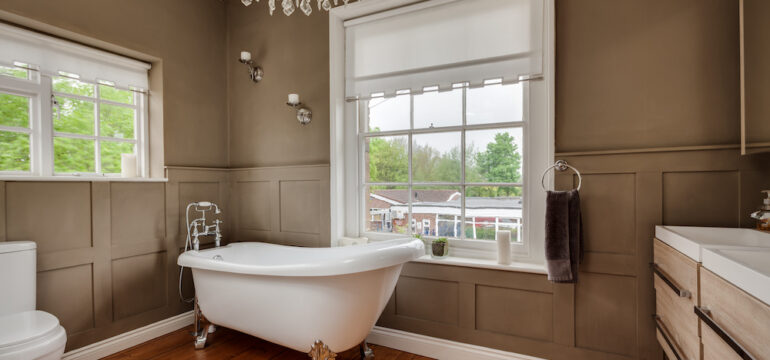 Traditional styled bathroom in an older home with roll top bath and half paneled walls within former old rectory with exposed timber floors, large chandelier, and windows to two aspects.