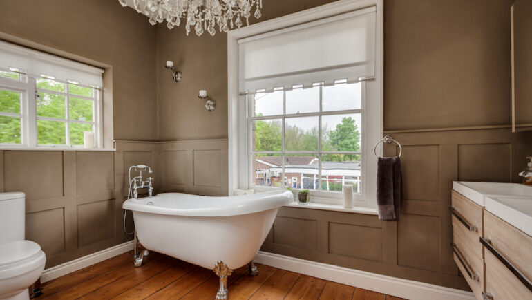 Traditional styled bathroom in an older home with roll top bath and half paneled walls within former old rectory with exposed timber floors, large chandelier, and windows to two aspects.