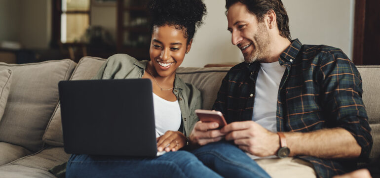 A happy young couple uses a laptop and cell phone to calculate their home equity while relaxing on a couch at home.