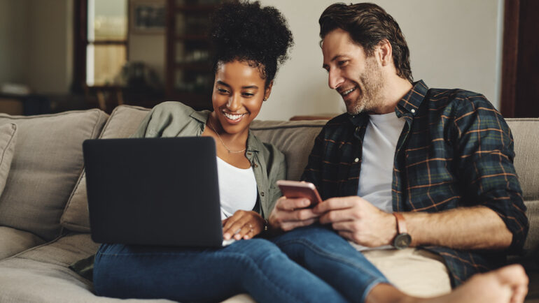 A happy young couple uses a laptop and cell phone to calculate their home equity while relaxing on a couch at home.