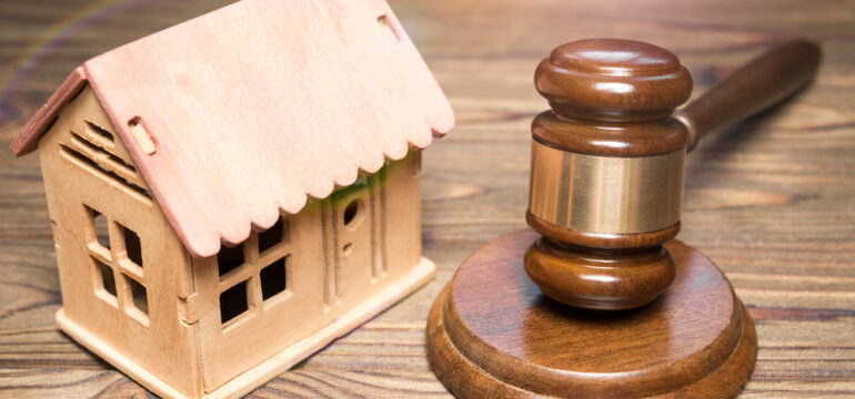 A hammer of a judge and a breadboard model of a wooden house against the background of a wooden table represent a home seizure under housing law.