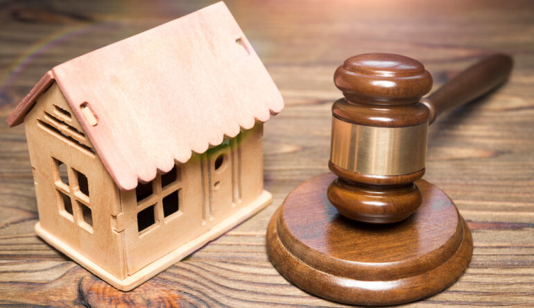 A hammer of a judge and a breadboard model of a wooden house against the background of a wooden table represent a home seizure under housing law.