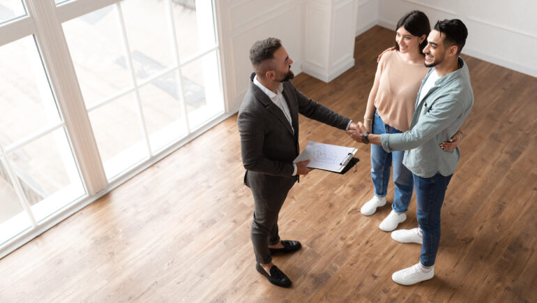 View of a millennial couple buying a new house, and shaking hands with a real estate agent. Concept shows importance of buyer agency agreements when buying a home.