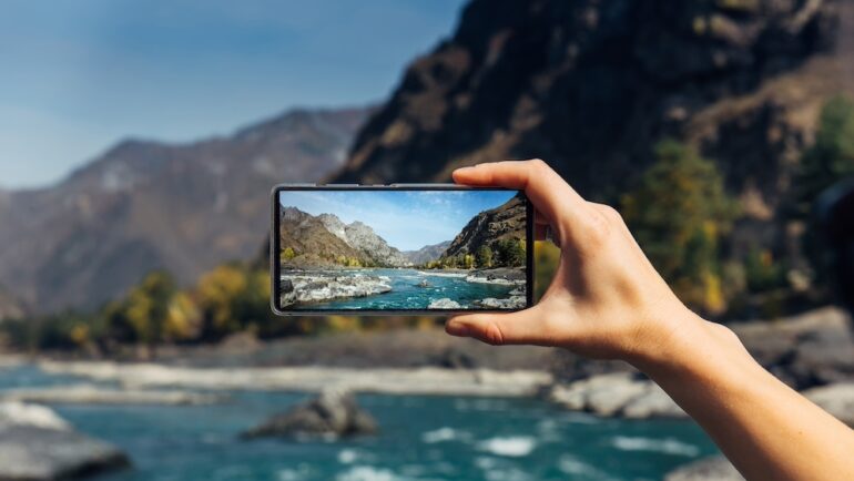 Female hand with smartphone close-up. Taking digital photo on smartphone while traveling. Delightful mountain landscape, stormy river, rocks and blue sky.