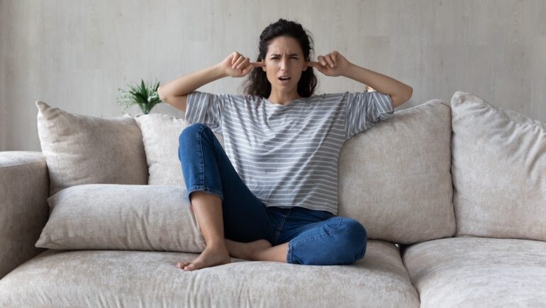 Unhappy woman plugging ears with fingers, sitting on couch at home. Annoying neighbor behaviors concept. Irritated young female ignoring avoiding loud sound of neighbors.