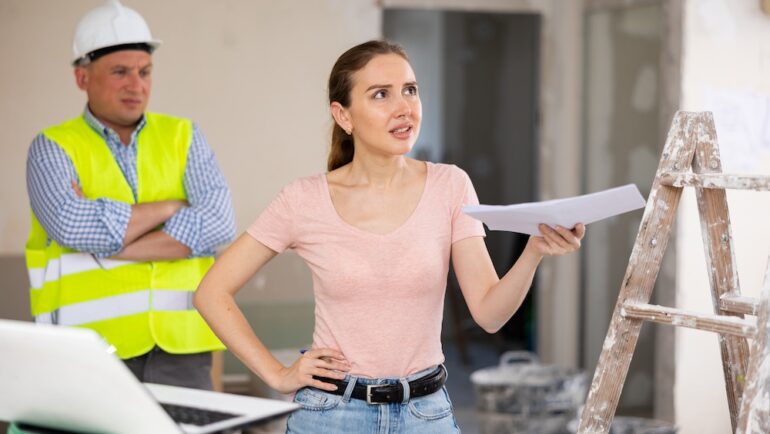 Frustrated young adult female designer arguing with male worker about subpar contractor work while examining indoor construction site.