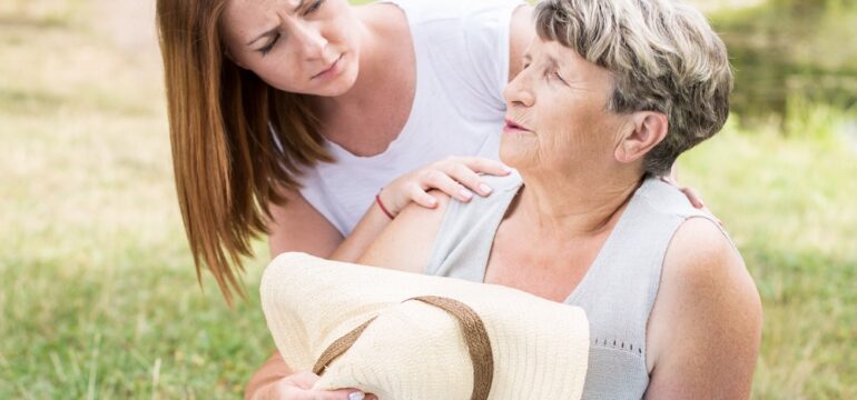 Image of an older woman sitting on the grass on a sunny day, feeling faint, and a young woman assisting her after working outside in the extreme heat.