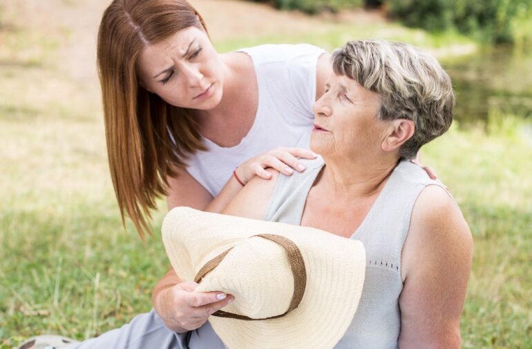 Image of an older woman sitting on the grass on a sunny day, feeling faint, and a young woman assisting her after working outside in the extreme heat.